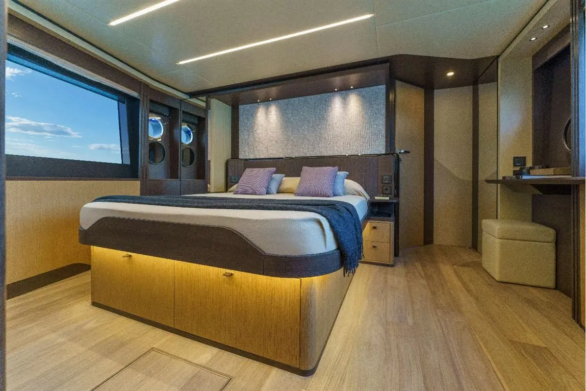 Modern yacht cabin featuring a double bed with purple cushions, ambient lighting, and large windows showcasing a blue sky.